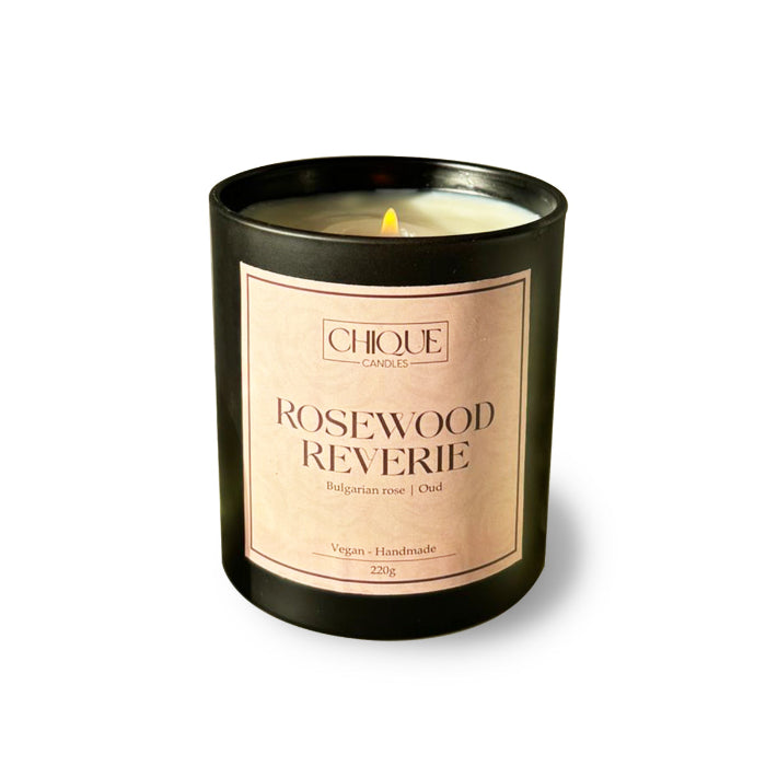 A black candle with a label that reads 'ROSEWOOD REVERIE' in a white font, indicating the scent or name of the product.