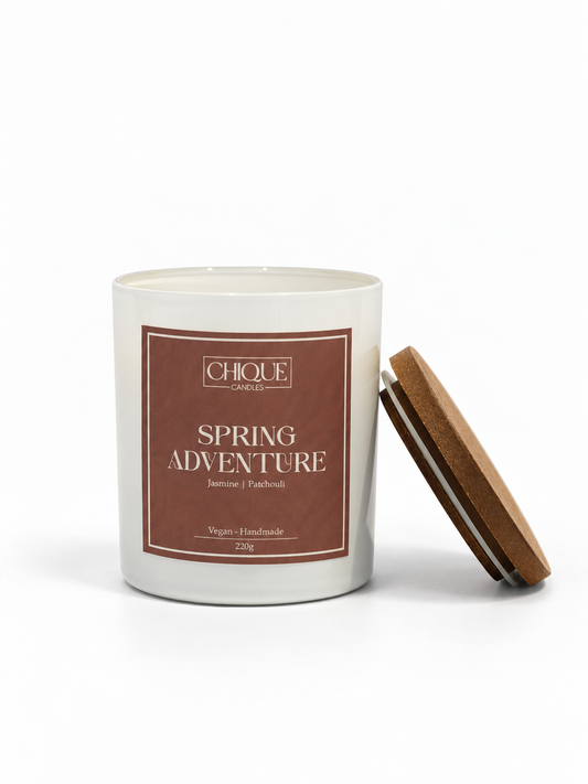 A white glass jar candle with a brown label reading 'CHIQUE CANDLESPRODUCTS SPRING ADVENTURE' and a matching brown lid leaning against the jar on a white background.
